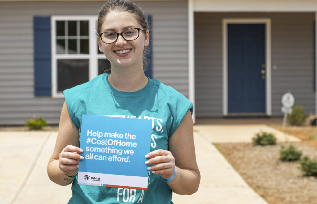 A smiling woman holding an advocacy statement card in front of a Habitat for Humanity house, "Helping make the Cost of Home something we can afford."